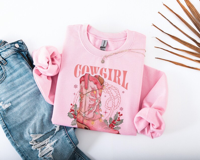 pink Sweatshirt with Western Christmas graphic design featuring a pink cowgirl boot wrapped in holiday lights, holly berries, rope lasso graphic, and text "Cowgirl Christmas"; festive Western holiday sweatshirt, country Christmas sweater, cowgirl aesthetic clothing, cozy winter pullover, Western Christmas gift, farmhouse holiday outfit, country girl Christmas apparel.
