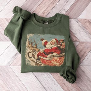 green Christmas sweatshirt with vintage 60s Santa Claus riding sleigh and reindeer illustration; retro Santa Christmas sweater, classic holiday sweatshirt, cozy winter pullover, nostalgic Christmas design, unisex festive apparel, holiday gift idea, traditional Christmas aesthetic sweatshirt, matching family Christmas outfits.