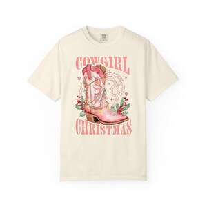 ivory comfort colors shirt with Western Christmas graphic design featuring a pink cowgirl boot wrapped in holiday lights, holly berries, rope lasso graphic, and text "Cowgirl Christmas"; festive Western holiday tee, country Christmas t-shirt, cowgirl aesthetic clothing, cozy winter sweatshirt, Western Christmas gift, farmhouse holiday outfit, country girl Christmas sweater.