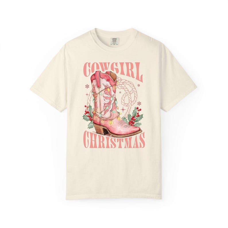 ivory comfort colors shirt with Western Christmas graphic design featuring a pink cowgirl boot wrapped in holiday lights, holly berries, rope lasso graphic, and text "Cowgirl Christmas"; festive Western holiday tee, country Christmas t-shirt, cowgirl aesthetic clothing, cozy winter sweatshirt, Western Christmas gift, farmhouse holiday outfit, country girl Christmas sweater.