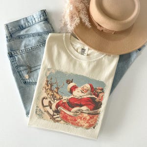 ivory Christmas comfort colors shirt with vintage 60s Santa Claus riding sleigh and reindeer illustration; retro Santa Christmas tee, classic holiday t-shirt, cozy winter top, nostalgic Christmas graphic design, unisex festive crewneck, holiday gift idea, traditional Christmas aesthetic sweatshirt, matching family Christmas outfits sweater.