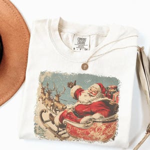 ivory Christmas comfort colors shirt with vintage 60s Santa Claus riding sleigh and reindeer illustration; retro Santa Christmas tee, classic holiday t-shirt, cozy winter top, nostalgic Christmas graphic design, unisex festive crewneck, holiday gift idea, traditional Christmas aesthetic sweatshirt, matching family Christmas outfits sweater.