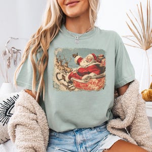 light-blue Christmas comfort colors shirt with vintage 60s Santa Claus riding sleigh and reindeer illustration; retro Santa Christmas tee, classic holiday t-shirt, cozy winter top, nostalgic Christmas graphic design, unisex festive crewneck, holiday gift idea, traditional Christmas aesthetic sweatshirt, matching family Christmas outfits sweater.