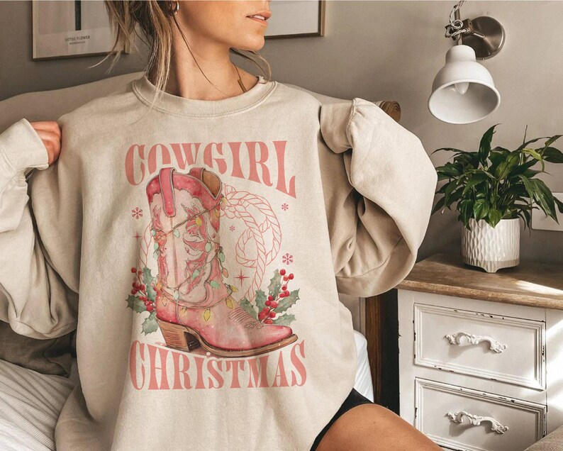 sand beige Sweatshirt with Western Christmas graphic design featuring a pink cowgirl boot wrapped in holiday lights, holly berries, rope lasso graphic, and text "Cowgirl Christmas"; festive Western holiday sweatshirt, country Christmas sweater, cowgirl aesthetic clothing, cozy winter pullover, Western Christmas gift, farmhouse holiday outfit, country girl Christmas apparel.