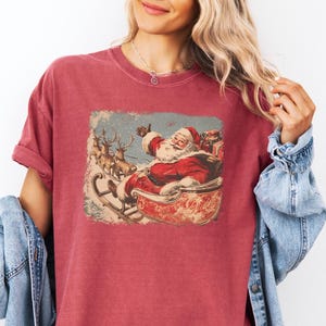 red crimson Christmas comfort colors shirt with vintage 60s Santa Claus riding sleigh and reindeer illustration; retro Santa Christmas tee, classic holiday t-shirt, cozy winter top, nostalgic Christmas graphic design, unisex festive crewneck, holiday gift idea, traditional Christmas aesthetic sweatshirt, matching family Christmas outfits sweater.