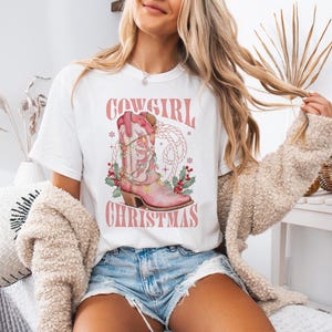 white comfort colors shirt with Western Christmas graphic design featuring a pink cowgirl boot wrapped in holiday lights, holly berries, rope lasso graphic, and text "Cowgirl Christmas"; festive Western holiday tee, country Christmas t-shirt, cowgirl aesthetic clothing, cozy winter sweatshirt, Western Christmas gift, farmhouse holiday outfit, country girl Christmas sweater.
