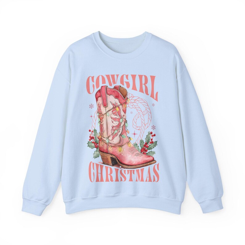 light blue Sweatshirt with Western Christmas graphic design featuring a pink cowgirl boot wrapped in holiday lights, holly berries, rope lasso graphic, and text "Cowgirl Christmas"; festive Western holiday sweatshirt, country Christmas sweater, cowgirl aesthetic clothing, cozy winter pullover, Western Christmas gift, farmhouse holiday outfit, country girl Christmas apparel.