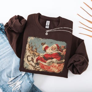 brown Christmas sweatshirt with vintage 60s Santa Claus riding sleigh and reindeer illustration; retro Santa Christmas sweater, classic holiday sweatshirt, cozy winter pullover, nostalgic Christmas design, unisex festive apparel, holiday gift idea, traditional Christmas aesthetic sweatshirt, matching family Christmas outfits.