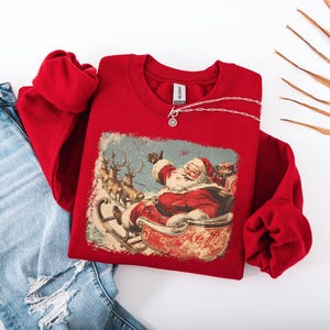 red Christmas sweatshirt with vintage 60s Santa Claus riding sleigh and reindeer illustration; retro Santa Christmas sweater, classic holiday sweatshirt, cozy winter pullover, nostalgic Christmas design, unisex festive apparel, holiday gift idea, traditional Christmas aesthetic sweatshirt, matching family Christmas outfits.