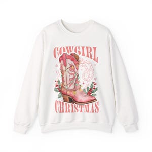 white Sweatshirt with Western Christmas graphic design featuring a pink cowgirl boot wrapped in holiday lights, holly berries, rope lasso graphic, and text "Cowgirl Christmas"; festive Western holiday sweatshirt, country Christmas sweater, cowgirl aesthetic clothing, cozy winter pullover, Western Christmas gift, farmhouse holiday outfit, country girl Christmas apparel.
