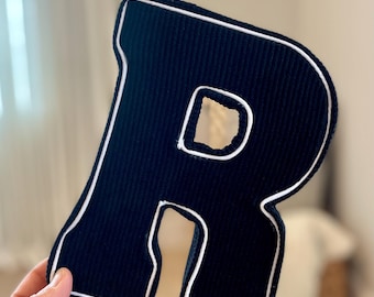 May include: A navy blue, corduroy letter "R" with white trim. The letter is a decorative item, possibly for a nursery or home decor. The white trim outlines the entire letter, including the inner cutout.