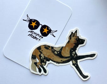 Peut inclure: Autocollant représentant un chien sauvage africain courant, avec un pelage tacheté de beige, noir et blanc. Une carte blanche avec des lunettes de soleil et le texte "Morques Mames" est en arrière-plan.