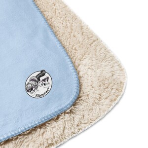 Pu&ograve; includere: Una coperta azzurra con una morbida fodera in sherpa color crema. La coperta presenta una toppa circolare con il disegno di due teschi che indossano occhiali da sole e la scritta Fibroeaz stampata in nero sulla superficie liscia azzurra.