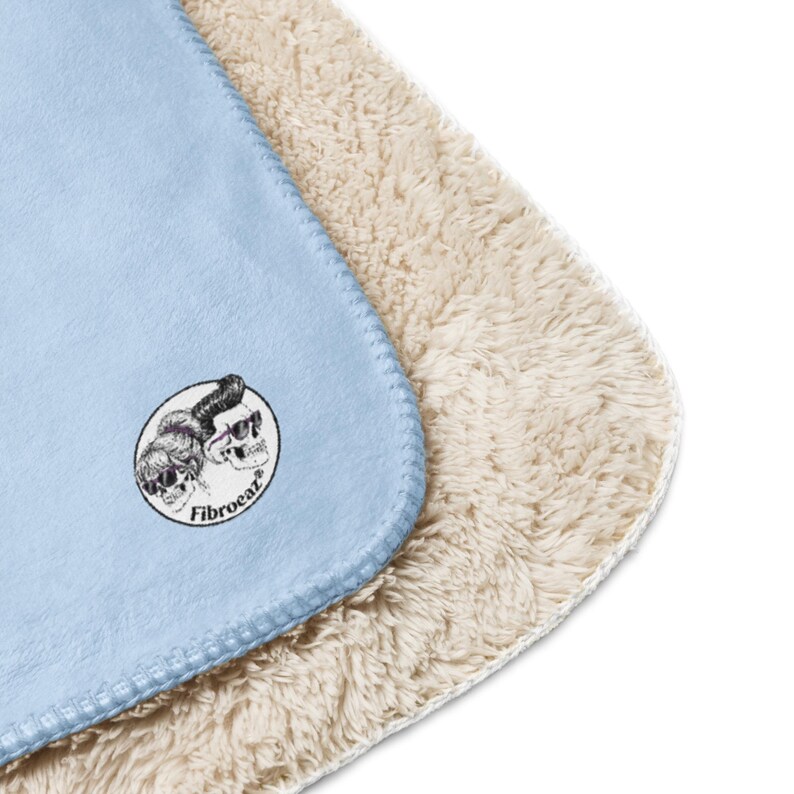 Pu&ograve; includere: Una coperta azzurra con una morbida fodera in sherpa color crema. La coperta presenta una toppa circolare con il disegno di due teschi che indossano occhiali da sole e la scritta Fibroeaz stampata in nero sulla superficie liscia azzurra.