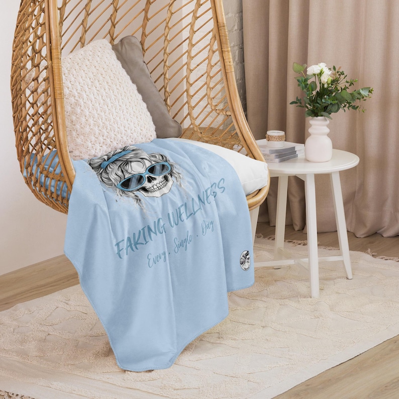 Pu&ograve; includere: Una coperta azzurra con un'illustrazione grafica di un teschio che indossa occhiali da sole blu e una fascia. Il testo "FAKING WELLNESS Every. Single. Day." &egrave; stampato in blu scuro sulla superficie del tessuto.