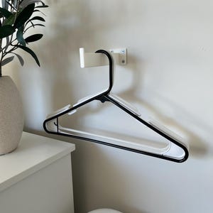 May include: A black and white clothes hanger mounted on a white wall. The hanger is attached to a white wall mount. The hanger is made of black metal and white plastic. A white cabinet and a plant are in the background.