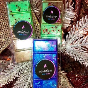 May include: Several wax melt bars in clear packaging. The bars are various colors, including green, blue, and gold, with glitter accents. Each bar has a circular label that reads "scentlylicious wax melts" with a flame logo. The background features festive decorations.