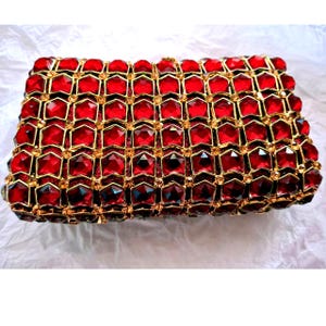 May include: A rectangular red clutch bag with a gold-coloured frame. The bag is covered in red, faceted gems, arranged in a honeycomb pattern. The gems are set in a gold-coloured metal frame, creating a luxurious design.