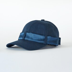 Op de afbeelding: Marineblauwe baseballcap met een satijnen band over de voorkant. De cap heeft een gebogen klep en een kleine knoop bovenop. De satijnen band is een iets donkerdere tint blauw.