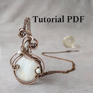 Op de afbeelding: Een handgemaakte koperdraad hanger met een grote, ronde, witte steen in het midden en kleine witte kralen. De hanger hangt aan een koperen ketting. De tekst "Tutorial PDF" staat in de afbeelding.