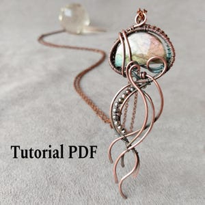 Puede incluir: Un collar con colgante artesanal de alambre de cobre con una piedra grande e iridiscente en el centro. El colgante presenta un intrincado enrollado de alambre y un diseño artístico. La cadena del collar es de color cobre a juego, y el texto "Tutorial PDF" es visible.