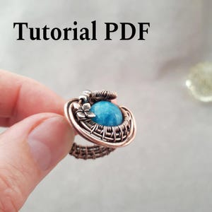 Könnte beinhalten: Handgefertigter Kupferdrahtring mit gewebtem Design und einem großen, runden, hellblauen Edelstein. Der Ring hat einen floralen Akzent, und der Text "Tutorial PDF" ist im Bild zu sehen.