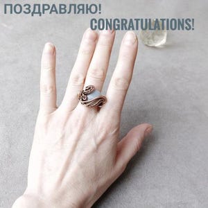Peut inclure: Une bague en fil de cuivre avec une pierre bleu clair est port&eacute;e sur une main. La bague pr&eacute;sente un design complexe. Les mots "F&eacute;licitations !" sont imprim&eacute;s en gris au-dessus de la main.