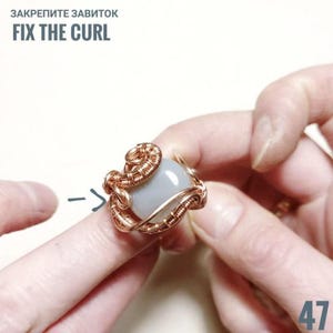 Peut inclure: Une bague en fil de cuivre avec une pierre bleu clair. La bague pr&eacute;sente un travail de filigrane complexe et le texte "FIX THE CURL". La bague est tenue par les doigts d'une personne.