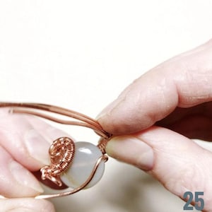 Peut inclure: Gros plan sur du fil de cuivre enroul&eacute; autour d'une perle de pierre grise claire. Deux mains manipulent le fil, formant un &eacute;l&eacute;ment d&eacute;coratif. Le projet artisanal est en cours, ax&eacute; sur la cr&eacute;ation de bijoux.