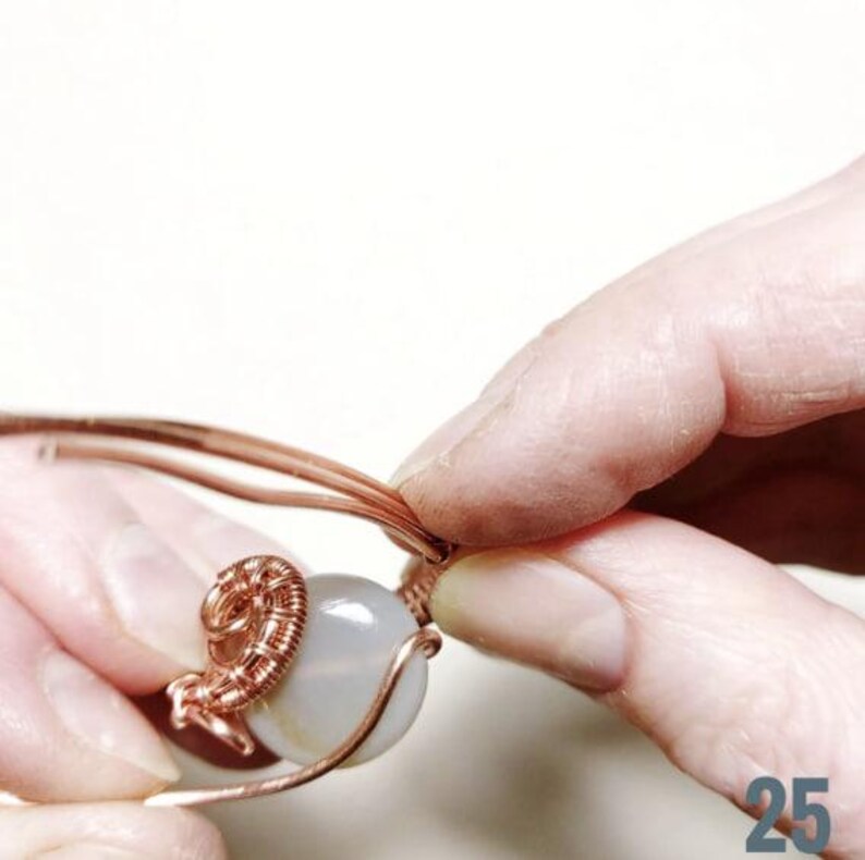 Peut inclure: Gros plan sur du fil de cuivre enroul&eacute; autour d'une perle de pierre grise claire. Deux mains manipulent le fil, formant un &eacute;l&eacute;ment d&eacute;coratif. Le projet artisanal est en cours, ax&eacute; sur la cr&eacute;ation de bijoux.
