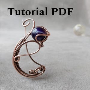 May include: A handcrafted copper wire ring with a dark blue, spherical gemstone. The wire is intricately wrapped around the stone and shaped into a unique, artistic design. The text "Tutorial PDF" is displayed at the top.