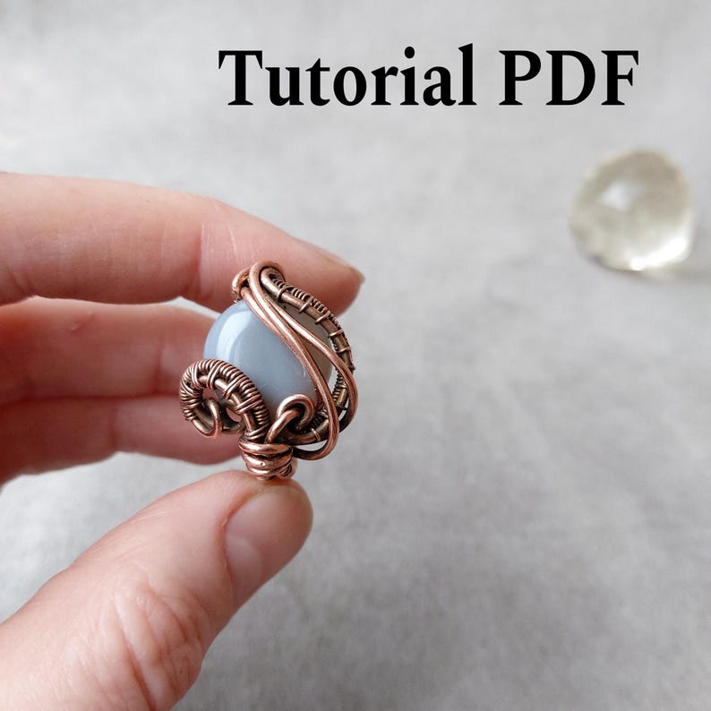 Peut inclure: Un pendentif artisanal enroul&eacute; de fil de cuivre, orn&eacute; d'une pierre bleu p&acirc;le. Le fil de cuivre est finement tiss&eacute; autour de la pierre, cr&eacute;ant un design artistique unique. Le texte "Tutorial PDF" est affich&eacute; en haut de l'image.