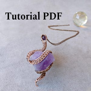 May include: A copper wire pendant necklace with a raw, purple amethyst crystal. The crystal is wrapped in copper wire, suspended from a delicate chain. The image includes the text "Tutorial PDF".