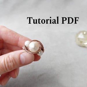 Könnte beinhalten: Handgefertigter Kupferdrahtring mit einer großen, runden, cremefarbenen Perle in der Mitte und kleineren Perlen an den Seiten. Der Ring wird in einer Hand gehalten. Der Text "Tutorial PDF" wird über dem Ring angezeigt.