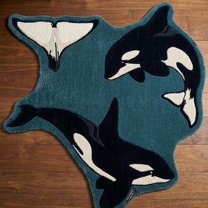 Puede incluir: Una alfombra de color verde azulado con forma irregular que presenta dos orcas negras y blancas y un diseño separado de aleta de cola de ballena. La alfombra tiene una superficie de pelo texturizado y una pequeña etiqueta de tela negra en el borde.