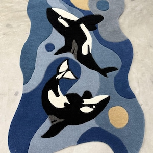 Puede incluir: Una alfombra de mechones con forma irregular que presenta dos orcas blancas y negras nadando entre patrones de agua abstractos en azul y azul claro, acentuada por formas circulares en tonos amarillos y grises suaves sobre la superficie texturizada.