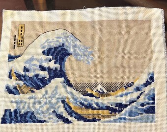 May include: Cross-stitch of Hokusai's "The Great Wave off Kanagawa." The artwork features a large wave in shades of blue, white, and yellow, with Mount Fuji in the background. The piece is on a beige canvas with Japanese text.