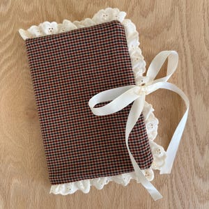May include: A small, rectangular fabric item with a red and black plaid pattern. The edges are trimmed with white eyelet lace. A white satin ribbon is tied in a bow on the side.