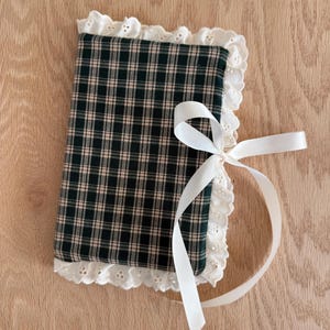 May include: A plaid fabric item with a white lace trim and a white ribbon tie. The fabric has a green and tan plaid pattern. The lace trim is scalloped. The ribbon is tied in a bow.