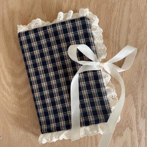 Puede incluir: Una funda de tela a cuadros azul marino y beige con un ribete de encaje blanco y una cinta color crema atada con un lazo. La funda, probablemente para un libro o diario, es rectangular. El fondo es una superficie de madera clara.