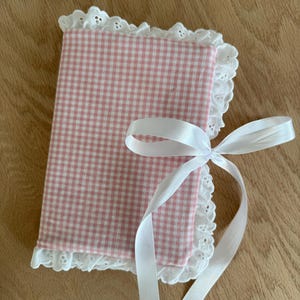 Puede incluir: Un cuaderno cubierto de tela de cuadros rosa y blanco, con un borde de encaje inglés blanco. Una cinta de raso blanca está atada con un lazo alrededor del cuaderno. El cuaderno está cerrado.