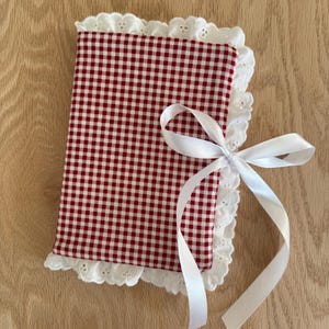 May include: A red and white gingham fabric bonnet with a white lace trim and a white satin ribbon bow. The bonnet is made of a soft fabric and is likely for a baby or child. The design is classic and evokes a vintage aesthetic.