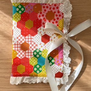 May include: A vibrant patchwork journal with a white lace trim and a white ribbon tie. The cover features a geometric pattern of hexagons, flowers, and polka dots in red, yellow, green, and pink. The journal is handmade and has a decorative edge.