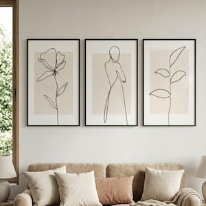 May include: Three framed art prints with black frames. Each print features a minimalist line drawing on a beige background. The drawings depict a flower, a human figure, and a plant. The artwork is displayed above a beige sofa with pillows.