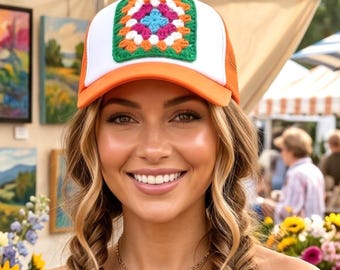 May include: A trucker hat featuring a white front panel with a colorful, square crochet granny square patch in shades of green, orange, pink, and blue. The hat has an orange brim and mesh back, paired with an orange off-the-shoulder long-sleeve top.
