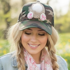 May include: A camouflage baseball cap with a white pom-pom and pink and white crochet bunny feet. The hat is a fun accessory for spring or Easter. The hat is made of a durable material and is a great gift idea.