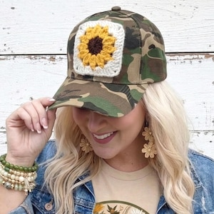 May include: Camouflage baseball cap with a square crochet patch featuring a yellow sunflower with a brown center. The cap is worn by a person wearing a denim jacket and a beige t-shirt with a floral design.