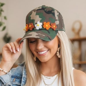 May include: Camouflage baseball cap adorned with crocheted flowers in shades of orange, white, and brown. The cap features a classic six-panel design and a curved brim. This accessory combines a military-inspired pattern with a touch of floral charm.