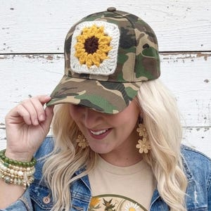 May include: A camouflage baseball cap featuring a square crochet patch on the front. The patch displays a yellow sunflower with a brown center on a white background. The cap has a structured crown and a curved brim, showcasing a classic design.
