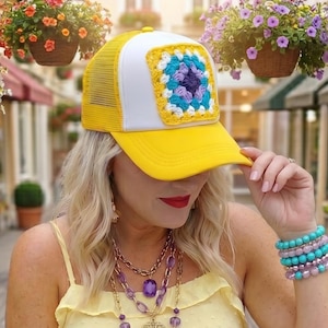 May include: A yellow and white trucker hat featuring a square crochet patch with blue, purple, and white yarn. The hat has a yellow brim and a white front panel with a yellow mesh back. The crochet design consists of concentric square layers.